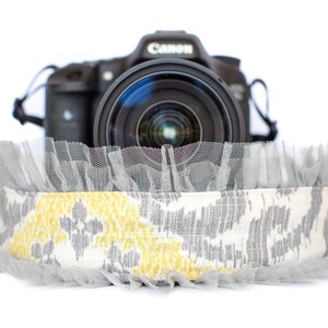 May include: A gray and yellow patterned camera strap with a ruffled edge, paired with a Canon camera. The strap features a repeating ikat-style design in gray and yellow on a white background, with a sheer gray ruffle.