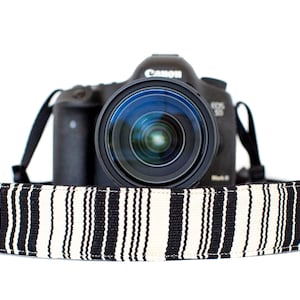 Puede incluir: Una cámara réflex digital Canon EOS 5D Mark III negra con una lente se muestra con una correa de cámara a rayas blancas y negras. La correa está hecha de un material tejido con rayas verticales. La lente refleja el entorno.