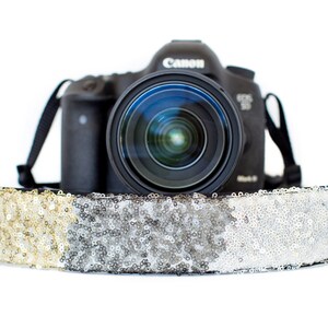 May include: A black Canon EOS 5D Mark III DSLR camera with a lens is resting on a decorative camera strap. The strap is covered in gold and silver sequins. The camera strap is a stylish accessory for photographers.