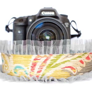May include: A black Canon camera rests atop a decorative band. The band features a colorful floral pattern in yellow, red, and blue, with a gray tulle trim. The camera lens is visible, suggesting photography or related accessories.