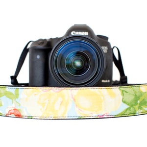 Puede incluir: Una cámara Canon EOS 6D Mark II negra con una correa floral. La correa tiene un fondo azul claro con flores amarillas, rosas y verdes. La lente de la cámara está centrada.