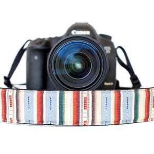 Puede incluir: Una cámara Canon DSLR negra con una lente se muestra con una colorida correa para cámara a rayas. La correa presenta rayas verticales en rojo, azul, crema y verde. La correa de la cámara mide aproximadamente 5 cm de ancho.