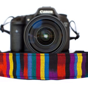 Puede incluir: Una cámara Canon negra con una lente grande, colocada sobre una correa de cámara con rayas verticales coloridas en rojo, amarillo, azul y morado. La correa destaca sobre un fondo blanco.