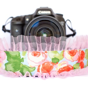 May include: A floral camera strap with a light blue background and red, pink, and green flowers. The strap is trimmed with pink tulle and sits in front of a Canon camera.