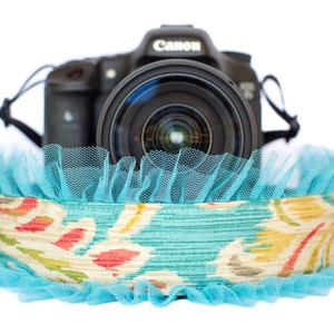 May include: A black Canon camera with a lens is positioned above a decorative band. The band features a floral pattern in teal, yellow, and red, trimmed with a ruffled turquoise net. The image is set against a white background.