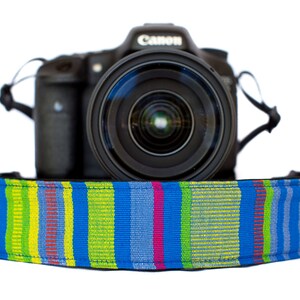 May include: A black Canon camera with a lens is shown with a colorful camera strap. The strap has a pattern of blue, green, yellow, and pink stripes. The camera is centered on a white background.