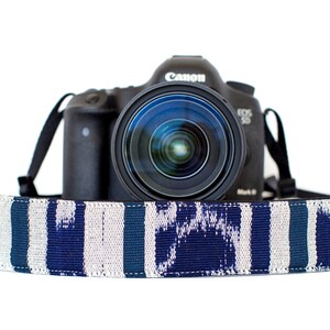 Puede incluir: Una cámara Canon EOS 5D Mark III negra con una lente se muestra, descansando sobre una correa de cámara estampada. La correa presenta un diseño ikat azul y blanco. El fondo es blanco.
