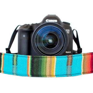 Puede incluir: Una cámara Canon negra con una colorida correa para cámara con estampado turquesa, rojo, naranja y verde. La correa está envuelta alrededor del cuerpo de la cámara, creando un contraste vibrante con la cámara negra.