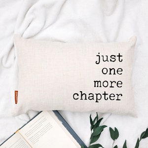 May include: A rectangular, beige pillow with the text "just one more chapter" in black lettering. An open book and green foliage are arranged below the pillow on a white surface, creating a cozy reading scene.