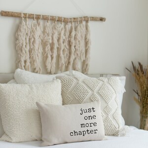 May include: A cozy bedroom scene featuring a bed with several white pillows of varying textures. A small rectangular pillow displays the text "just one more chapter." Above the bed hangs a decorative wall hanging made of cream-colored yarn.