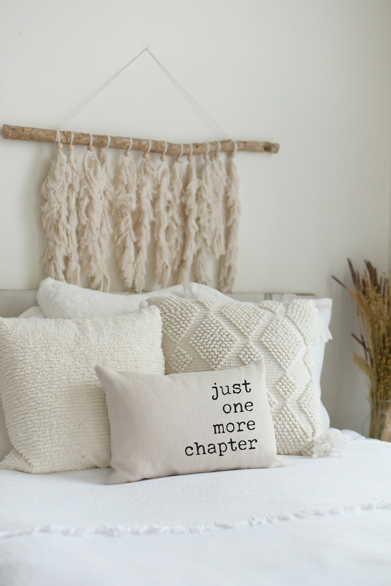 May include: A cozy bedroom scene featuring a bed with several white pillows of varying textures. A small rectangular pillow displays the text "just one more chapter." Above the bed hangs a decorative wall hanging made of cream-colored yarn.