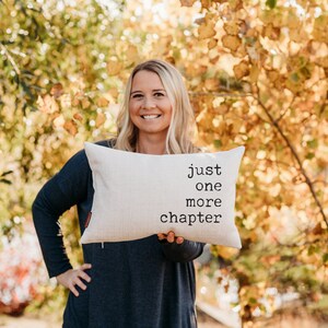 May include: Cream-colored rectangular pillow with the text "just one more chapter" in black. The pillow is held by a person in front of a blurred background of fall foliage.