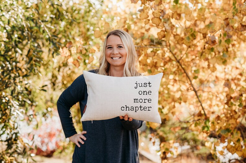 May include: Cream-colored rectangular pillow with the text "just one more chapter" in black. The pillow is held by a person in front of a blurred background of fall foliage.