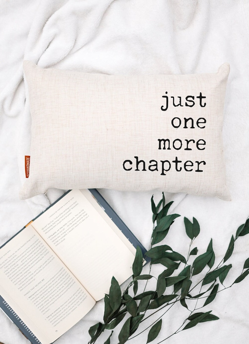 May include: A rectangular, beige pillow with the text "just one more chapter" in black lettering. An open book and green foliage are arranged below the pillow on a white surface, creating a cozy reading scene.