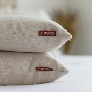 May include: Two beige linen throw pillows stacked on top of each other. Each pillow has a rectangular, brown tag that reads "521HANDMADE". The pillows are on a white surface.