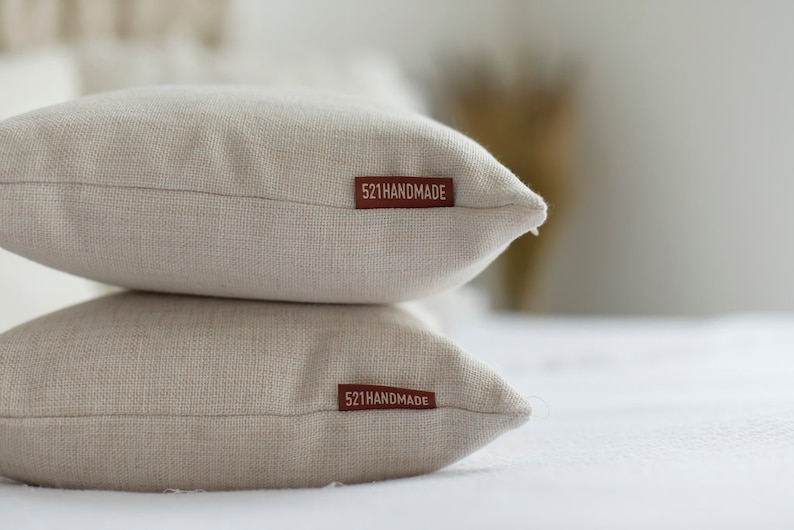 May include: Two beige linen throw pillows stacked on top of each other. Each pillow has a rectangular, brown tag that reads "521HANDMADE". The pillows are on a white surface.