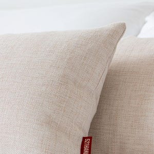 May include: Two beige linen throw pillows with a textured weave, resting on a white surface. A red tag on one pillow reads "521HANDMADE" in white text. The pillows have a soft, neutral color.