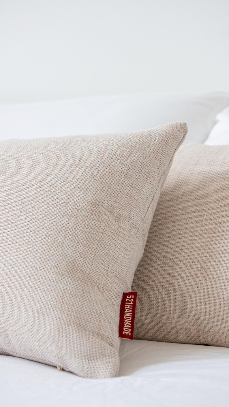 May include: Two beige linen throw pillows with a textured weave, resting on a white surface. A red tag on one pillow reads "521HANDMADE" in white text. The pillows have a soft, neutral color.