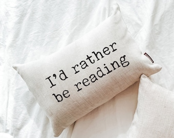 May include: A beige throw pillow with the text "I'd rather be reading" printed in black.