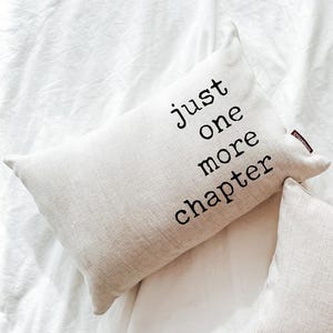 May include: A rectangular beige pillow with the text "just one more chapter" in black. The pillow is on a white sheet. The pillow has a small red tag on the side.