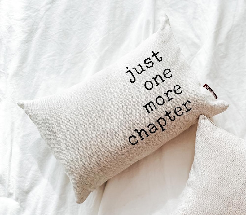 May include: A rectangular beige pillow with the text "just one more chapter" in black. The pillow is on a white sheet. The pillow has a small red tag on the side.