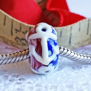Pode incluir: Uma conta de vidro vermelha, branca e azul com um design de &acirc;ncora branca. A conta est&aacute; em uma corrente prateada.