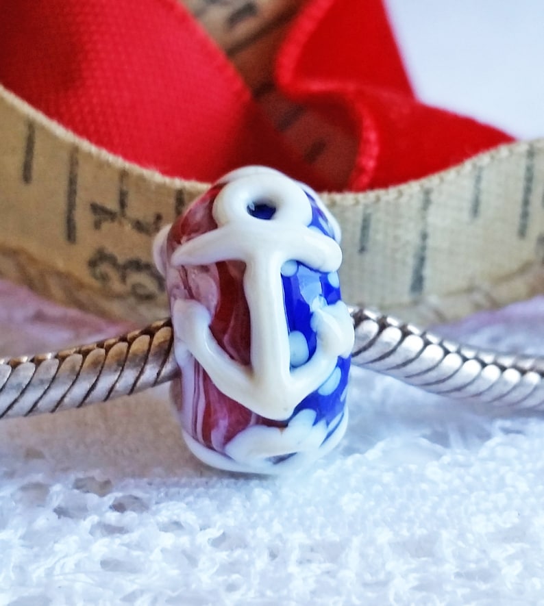 Pode incluir: Uma conta de vidro vermelha, branca e azul com um design de &acirc;ncora branca. A conta est&aacute; em uma corrente prateada.