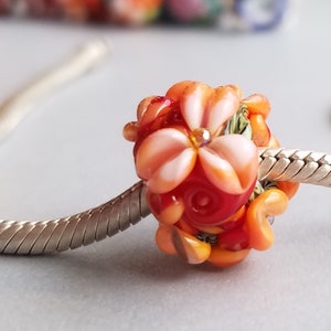 Pode incluir: Uma conta de vidro com um design de flor vermelha, laranja e branca. A conta está em uma corrente de prata.