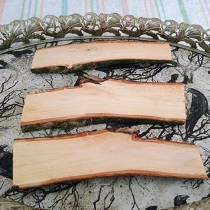 May include: Three rectangular pieces of light-colored wood with bark edges are arranged on a decorative tray. The tray has a silver-colored, ornate rim and a black and white patterned surface with crows and branches.