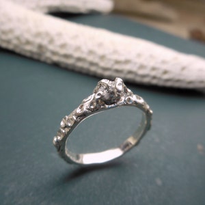 May include: A silver ring with a textured, organic design. The ring features a single, small, clear gemstone.