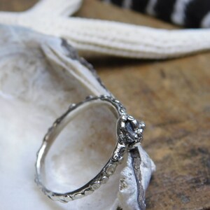 May include: A silver ring with a textured band and a rough, gray gemstone set in the center. The ring is resting on a white seashell.