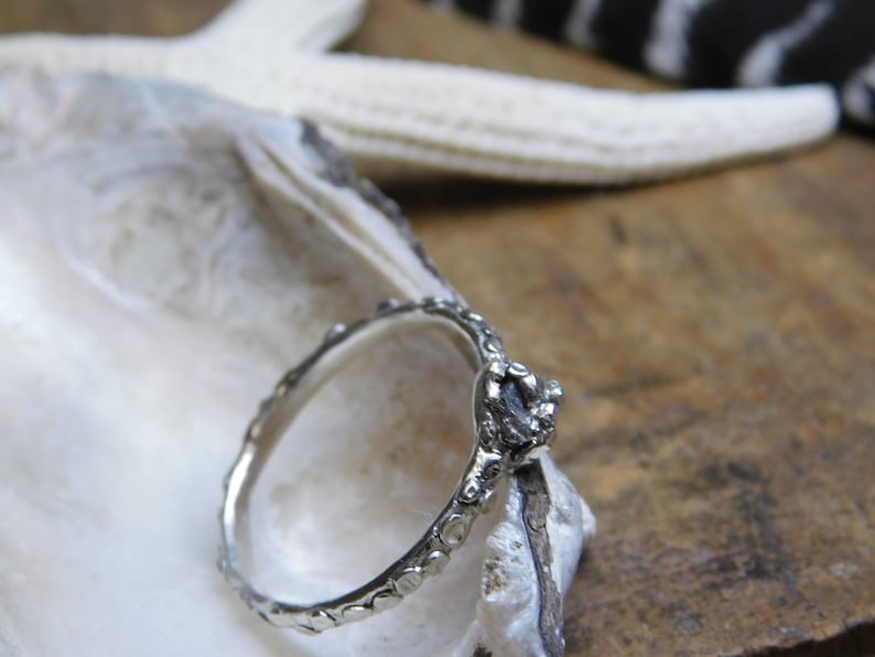 May include: A silver ring with a textured band and a rough, gray gemstone set in the center. The ring is resting on a white seashell.