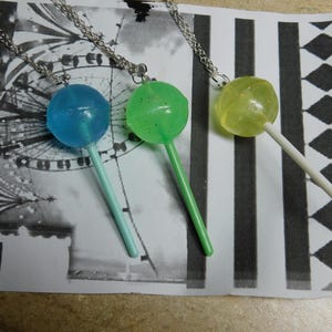 Puede incluir: Tres collares con colgantes de piruletas. Cada collar presenta un dije de piruleta de diferente color: azul, verde y amarillo. Los dijes están sujetos a cadenas de color plateado. Las piruletas están sobre un palito blanco.