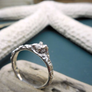 May include: A silver ring with a rough, textured band and a small, clear stone set in the center. The ring is sitting on a piece of driftwood.