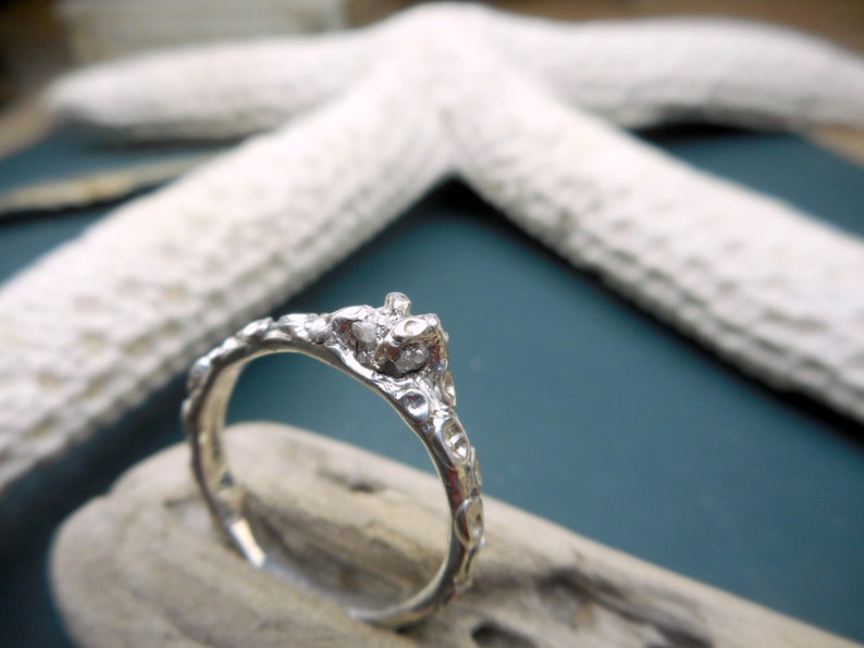 May include: A silver ring with a rough, textured band and a small, clear stone set in the center. The ring is sitting on a piece of driftwood.