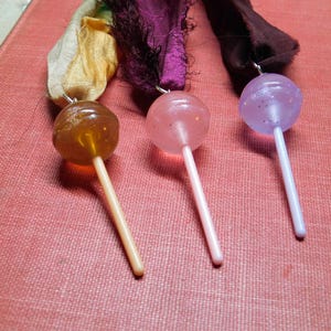 Puede incluir: Tres piruletas con esferas de caramelo claras y coloridas y palitos blancos. Las piruletas son ámbar, rosa y morado, cada una con una cinta decorativa. Las piruletas descansan sobre una superficie roja texturizada.