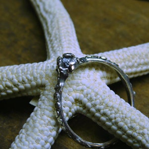 May include: A silver ring with a single, clear gemstone set in a textured band. The ring is displayed on a white starfish.
