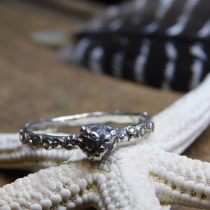 May include: A silver ring with a rough, dark gray stone set in the center. The band is decorated with a textured, organic design.
