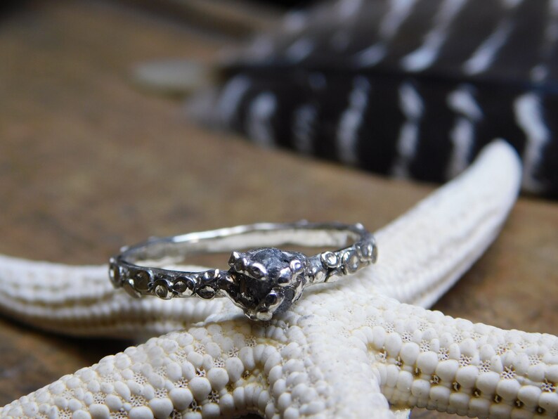 May include: A silver ring with a rough, dark gray stone set in the center. The band is decorated with a textured, organic design.