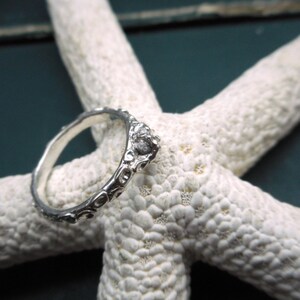 May include: A silver ring with a textured design, featuring a small, clear stone set in the center. The ring is displayed on a white starfish.