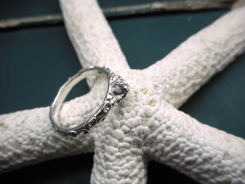 May include: A silver ring with a textured design, featuring a small, clear stone set in the center. The ring is displayed on a white starfish.