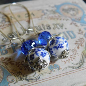 May include: Earrings featuring white porcelain beads with blue floral patterns, accented by faceted blue crystal beads. The earrings have silver-tone ear wires and decorative metal caps. The background is a vintage-style paper with text.