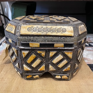 May include: A decorative wooden box with intricate carvings and inlaid bone accents. The box has a square shape with a raised lid and a base with a geometric pattern.