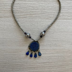 Puede incluir: Un collar de plata con un colgante de lapislázuli azul y cuentas de plata. El colgante es redondo con un diseño de lágrima.