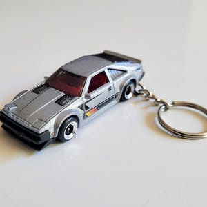 May include: A silver toy car keyring featuring a wedge-shaped sports car design with pop-up headlights, black trim, and red interior seating. The vehicle has a metallic finish, black wheels, and a silver metal split ring attached to the rear.