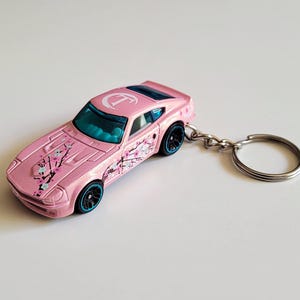 Puede incluir: Un llavero de coche de juguete rosa con una letra T blanca en el techo y un gráfico de rama de cerezo en el capó y el lateral. El vehículo tiene ventanas tintadas de azul, ruedas negras con llantas azules y una anilla de metal plateado.