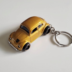 May include: A gold-coloured die-cast metal keyring in the shape of a classic car. The car has black wheels and a silver key ring. The car has a small emblem on the front. The keyring is on a white surface.