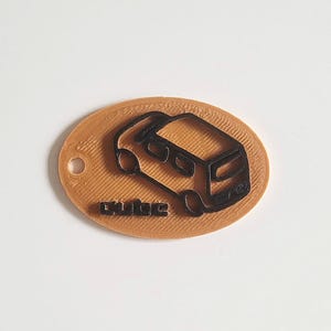 May include: A brown oval keyring featuring a black outline of a camper van. The word "dube" is printed in black at the bottom. The keyring has a small hole at the top.