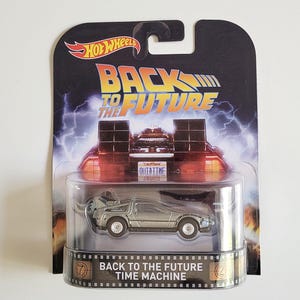 May include: A Hot Wheels die-cast model of the DeLorean time machine from the film "Back to the Future." The silver car has black wheels and is packaged in a clear plastic case with a themed backing card featuring the film's logo and imagery.