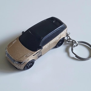 Könnte beinhalten: Ein goldfarbener Spielzeugauto-Schlüsselanhänger mit schwarzem Dach und schwarzen Rädern. Der Schlüsselanhänger hat einen silbernen Metallring. Das Auto hat die Nummer "68" an der Seite.